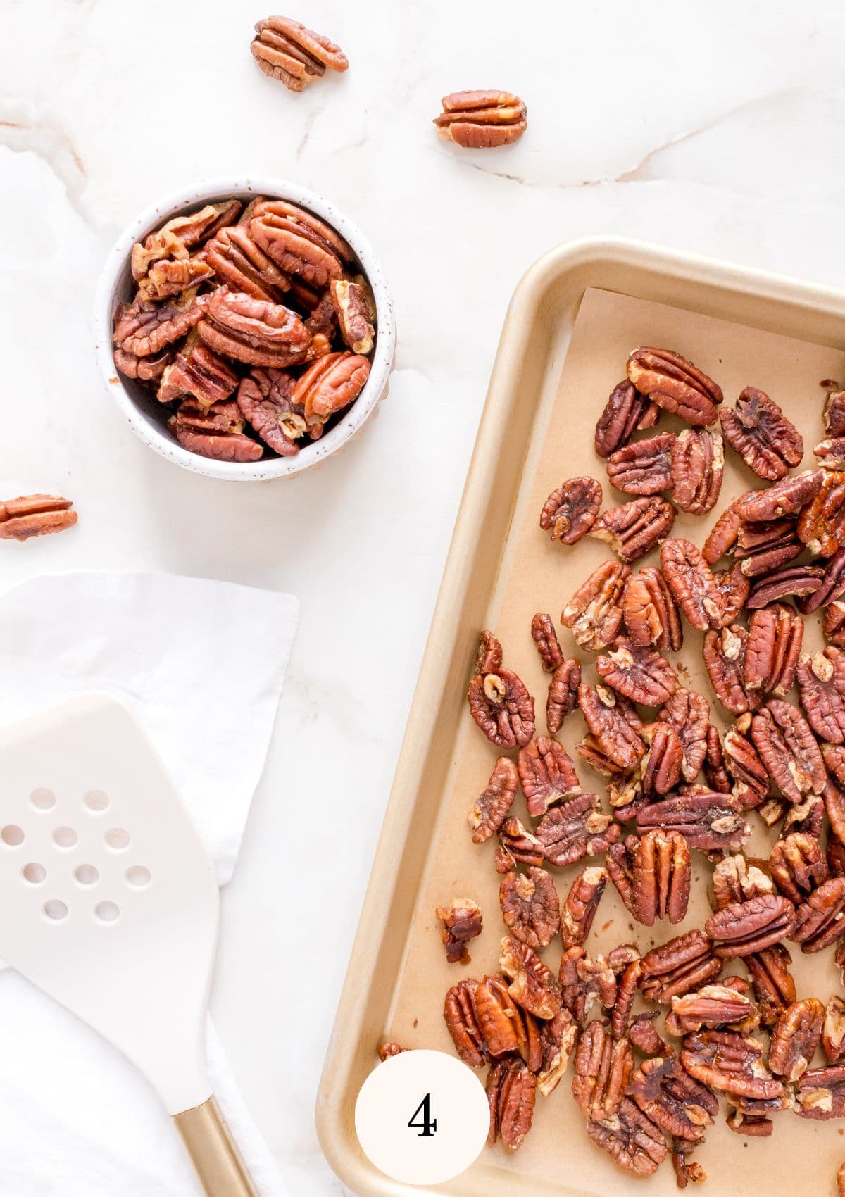 maple roasted pecans on gold baking sheet with a white spatula and white bowl filled with pecans