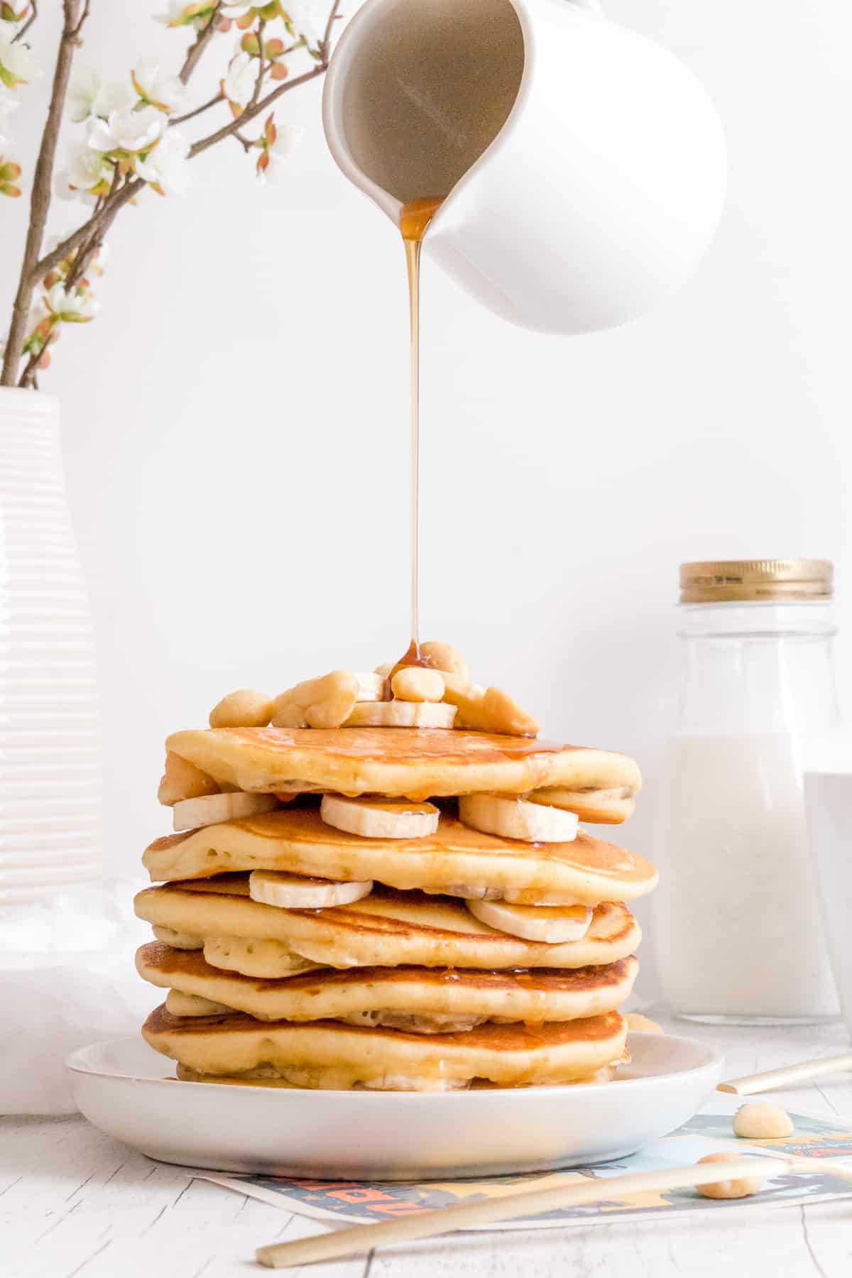 pouring maple syrup over a stack of banana macadamia nut pancakes.