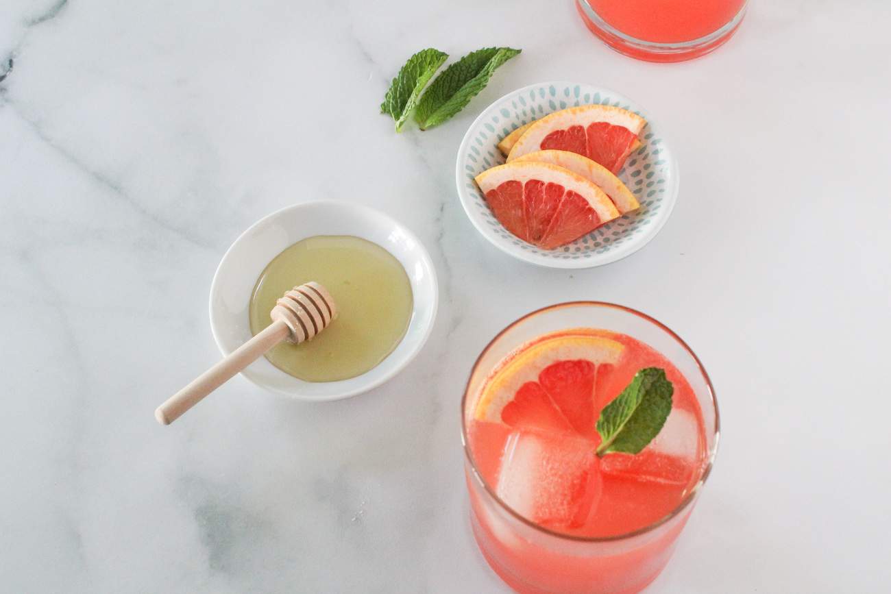 Sparkling Ruby Red Grapefruit Cocktail next to a bowl of honey and a bowl of sliced grapefruit