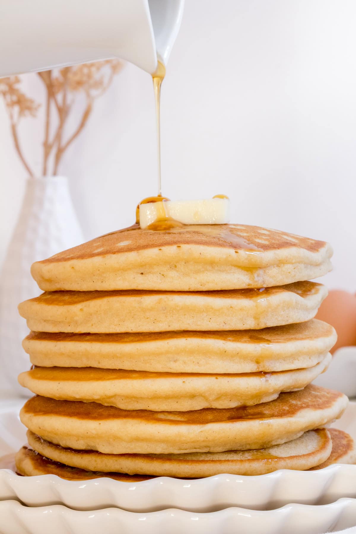 a white pitcher of syrup pouring over a stack of gluten free pancakes on a white plate.
