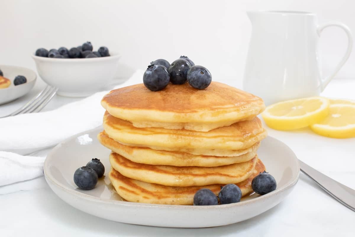 light and fluffy lemon pancakes on white plate with bowl of berries and pitcher of maple syrup