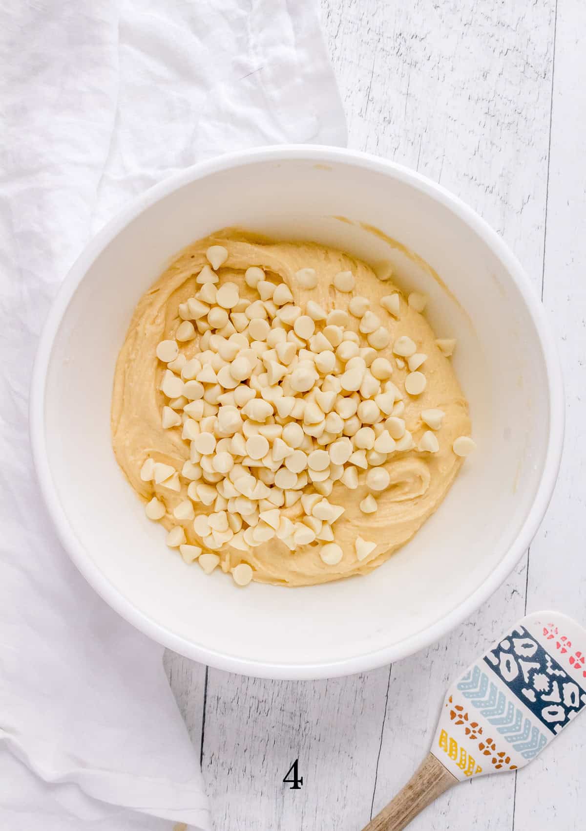 adding white chocolate chip to cookie dough in white mixing bowl. 