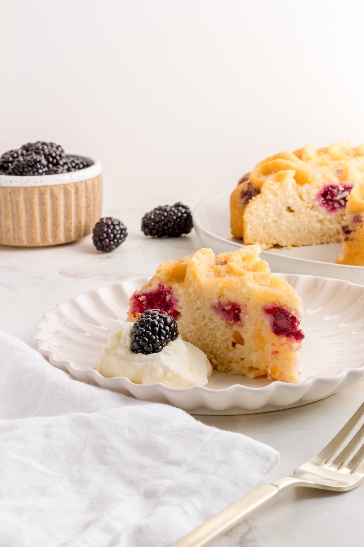 A slice of berry cake with fresh whipped cream topped with a blackberry sits on a white plate. In the background, a bowl of blackberries and the rest of the cake are visible. A fork and napkin are nearby.