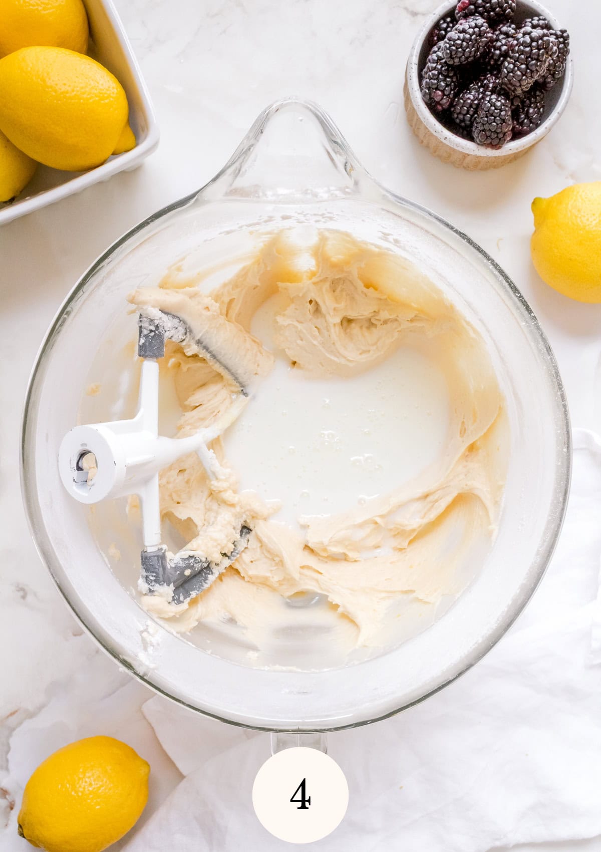 A glass mixing bowl batter, buttermilk and a paddle attachment, surrounded by fresh lemons and a small bowl of blackberries on a marble surface.