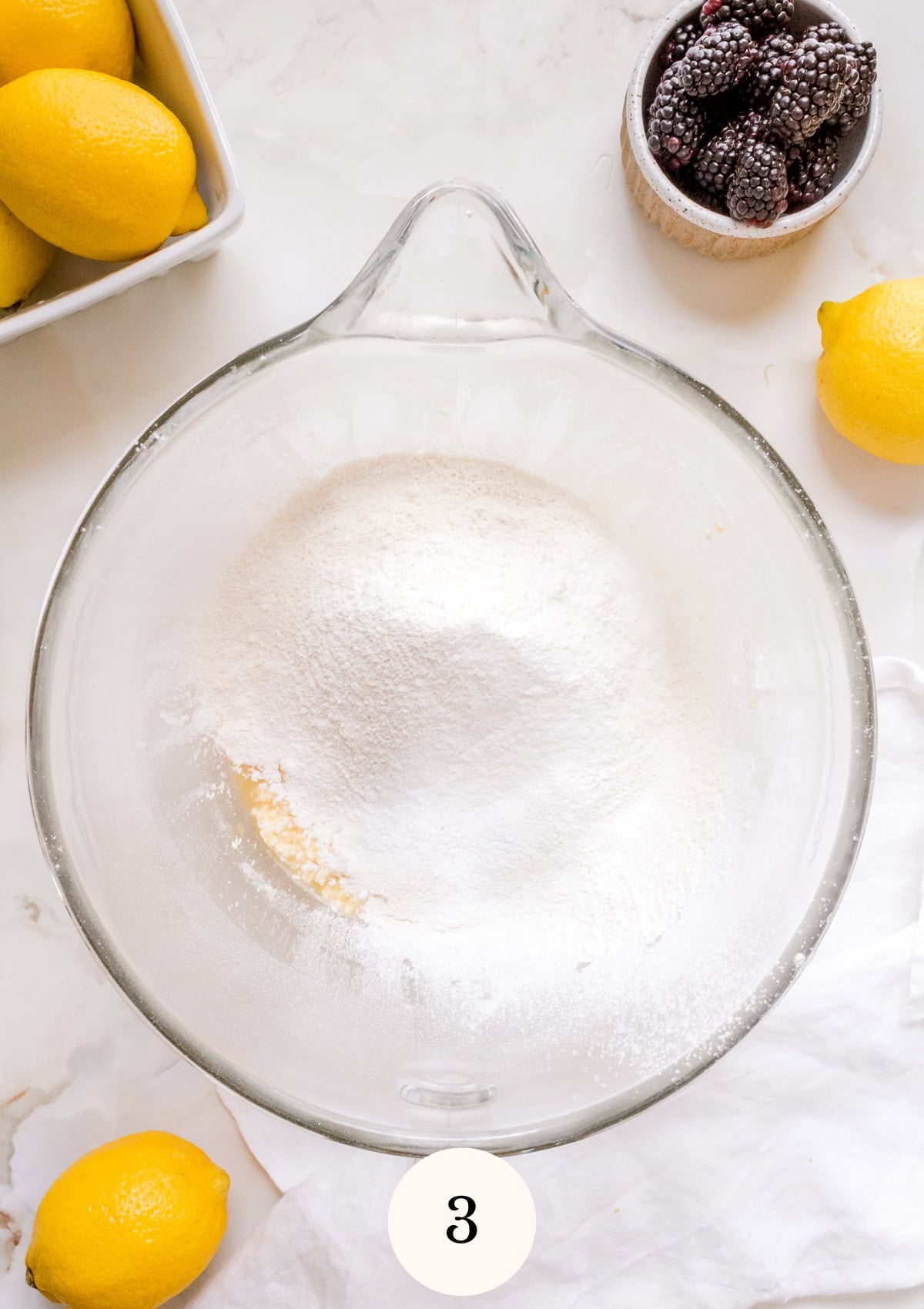 A glass mixing bowl with flour on top of creamed ingredients sits on a white surface, surrounded by fresh lemons, a small bowl of blackberries, and a white cloth.