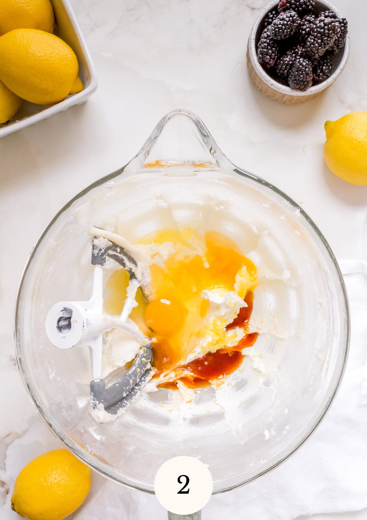 A glass mixing bowl with eggs, vanilla, and lemon zest being mixed. Surrounding the bowl are lemons, a small bowl of blackberries, and a white cloth. A 2 is displayed at the bottom center.