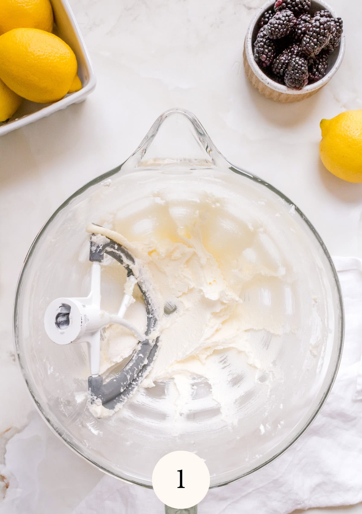Overhead view of a glass mixing bowl with a paddle attachment and creamed butter and sugar, surrounded by fresh lemons and a small bowl of blackberries on a marble countertop.