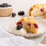 A slice of blackberry cake with visible blackberry pieces sits on a white plate next to a dollop of whipped cream topped with a fresh blackberry; additional cake and berries appear in the background.