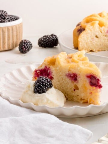 A slice of blackberry cake with visible blackberry pieces sits on a white plate next to a dollop of whipped cream topped with a fresh blackberry; additional cake and berries appear in the background.
