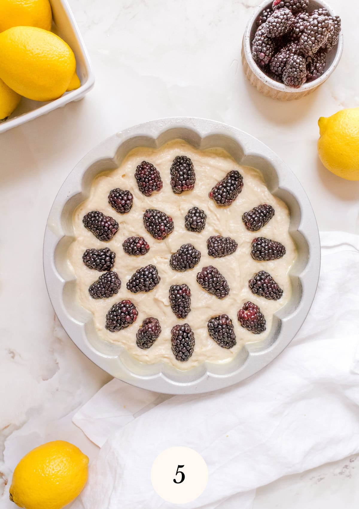 Round cake pan filled with unbaked batter topped with fresh blackberries, surrounded by lemons and a bowl of blackberries on a white marble surface.
