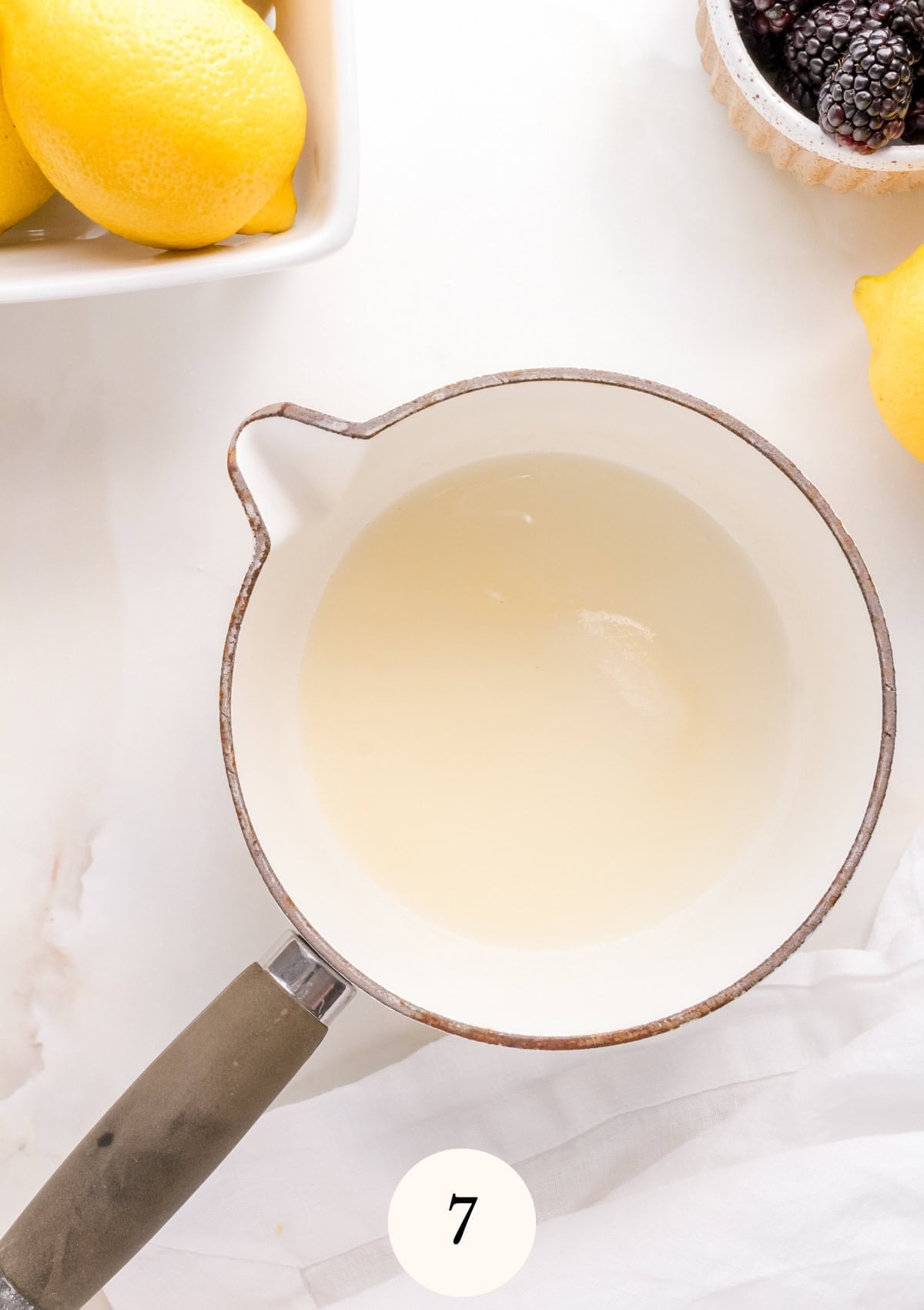 A saucepan filled with lemon juice and sugar sits on a white surface. Lemons are in a bowl nearby, and a small dish of blackberries.
