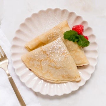 Two folded gluten free crepes dusted with powdered sugar on a scalloped white plate, garnished with two raspberries and a sprig of mint. A gold fork and a white napkin are placed beside the plate.