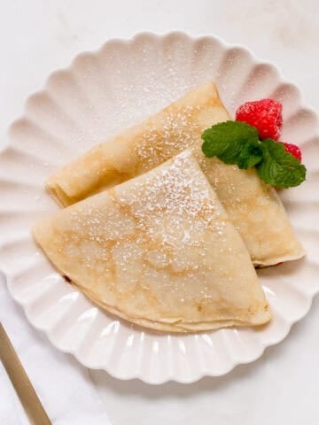 Two folded gluten free crepes dusted with powdered sugar on a scalloped white plate, garnished with two raspberries and a sprig of mint. A gold fork and a white napkin are placed beside the plate.