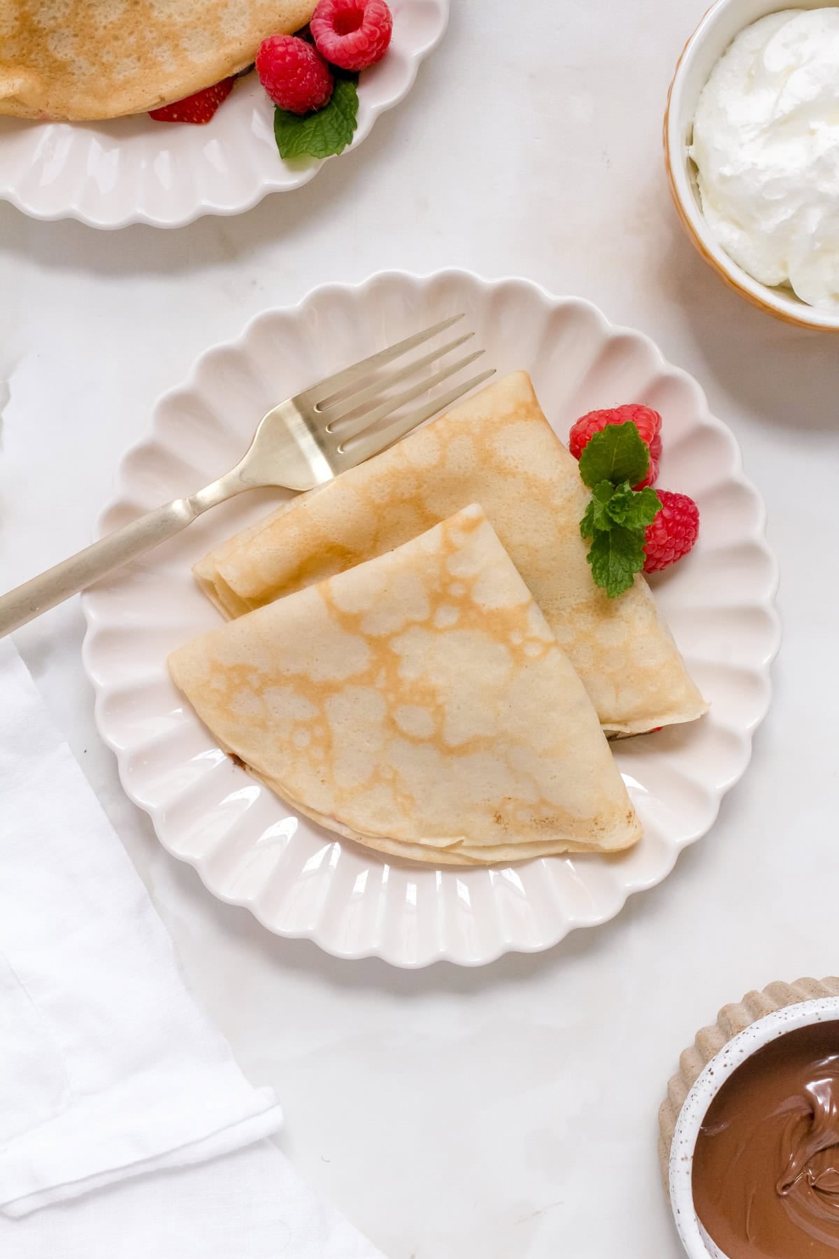 A plate with two folded gluten free crepes, garnished with fresh raspberries and mint, sits on a white surface next to a gold fork. A bowl of whipped cream and a small dish of Nutella spread are nearby.
