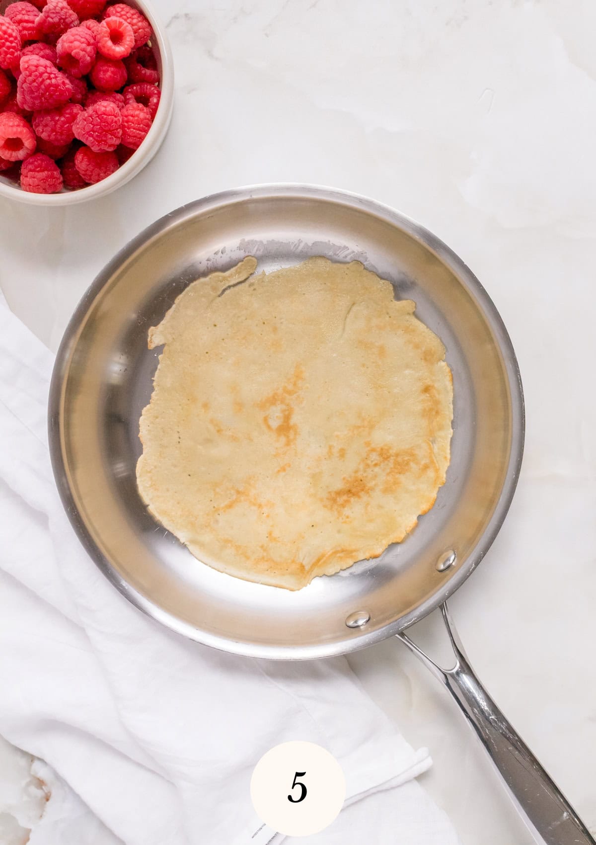A gluten free crepe cooking in a stainless steel pan on a white surface.