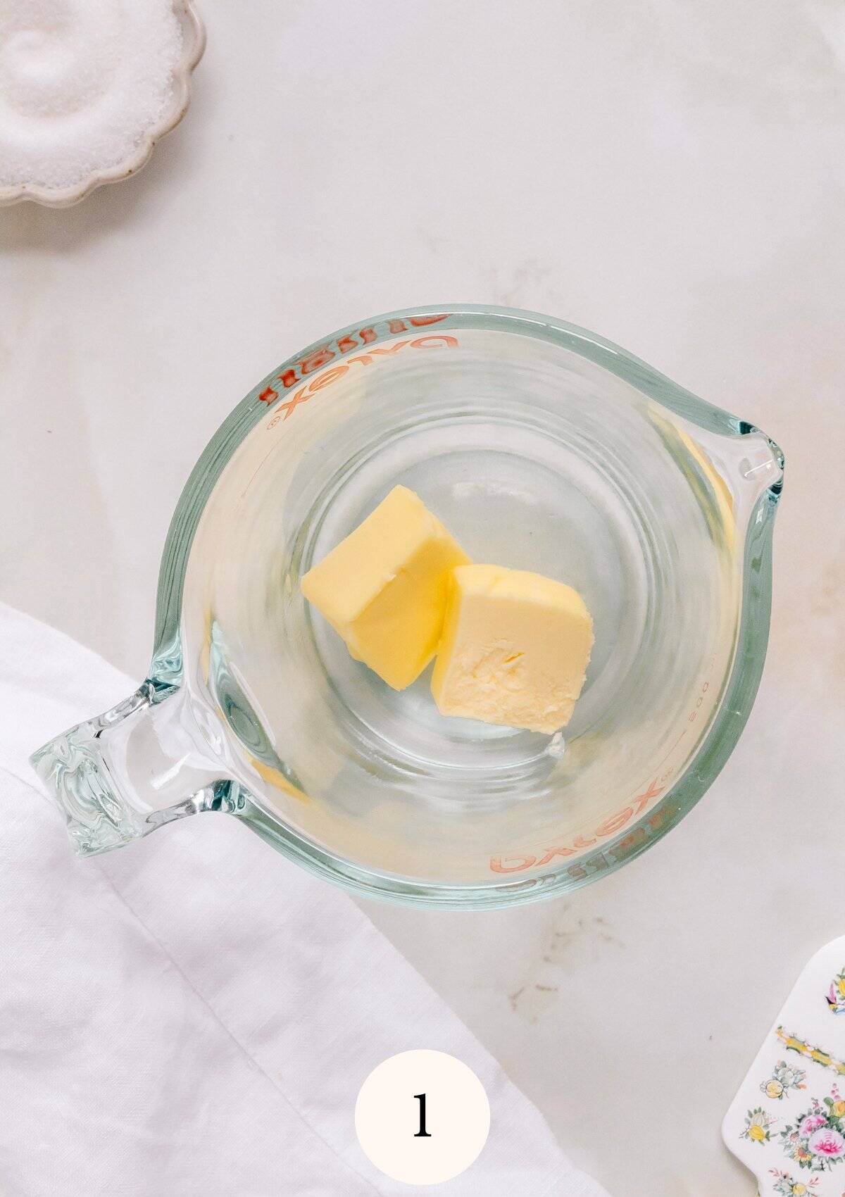 Overhead view of two pieces of butter in a glass measuring cup on a light countertop, ready to melt for gluten free crepes.
