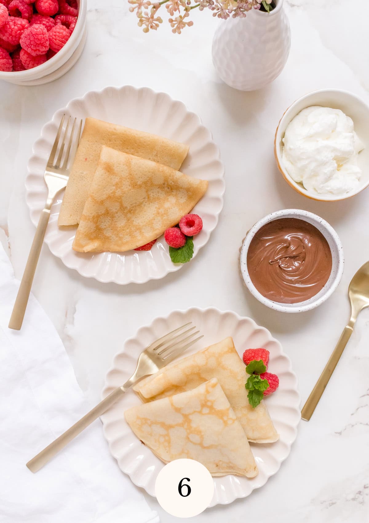 Two plates with folded gluten free crepes, fresh raspberries, and mint sprigs are set with gold forks. Nearby are bowls of raspberries, whipped cream, and chocolate spread on a white marble surface.