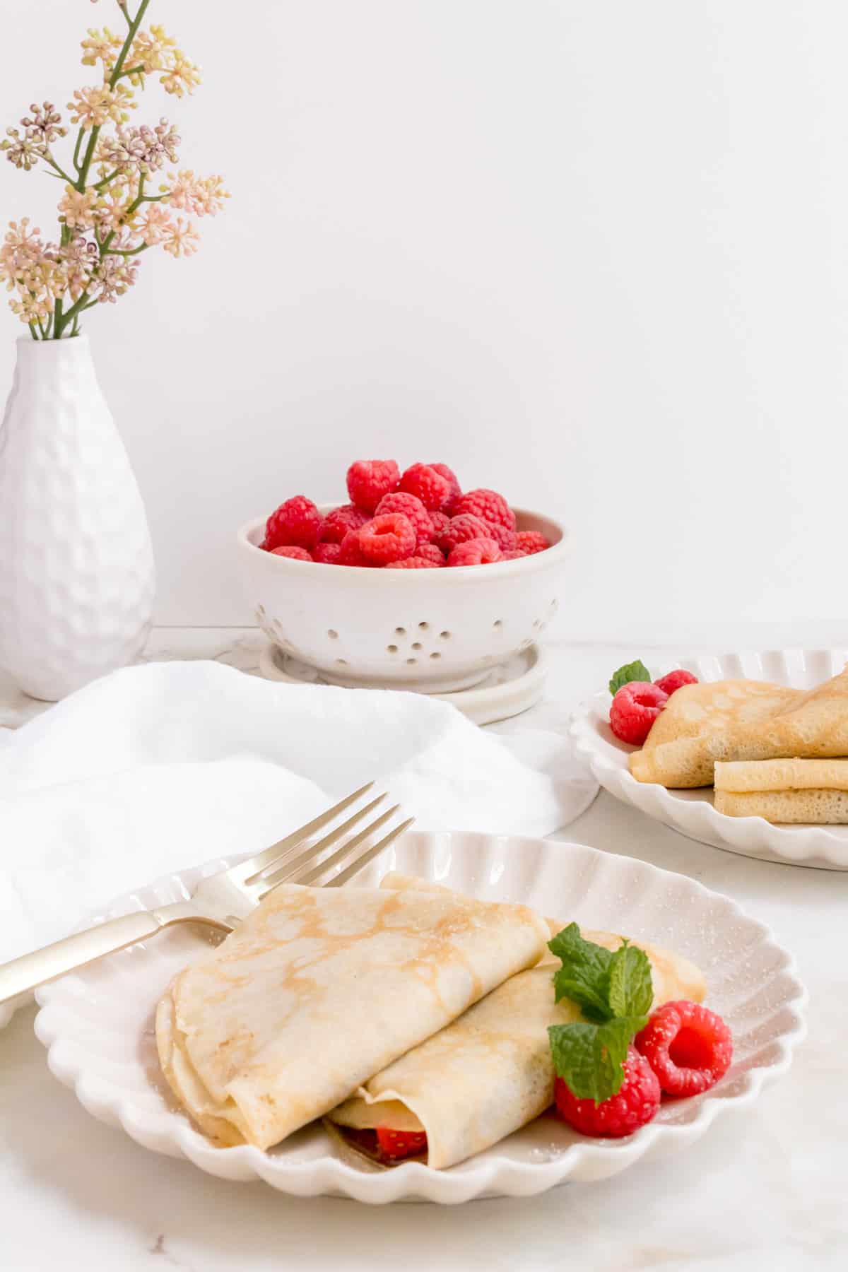 A plate of gluten free crepes garnished with raspberries and mint sits next to a fork, with a bowl of fresh raspberries, another plate of crepes, a white napkin, and a vase of flowers in the background.