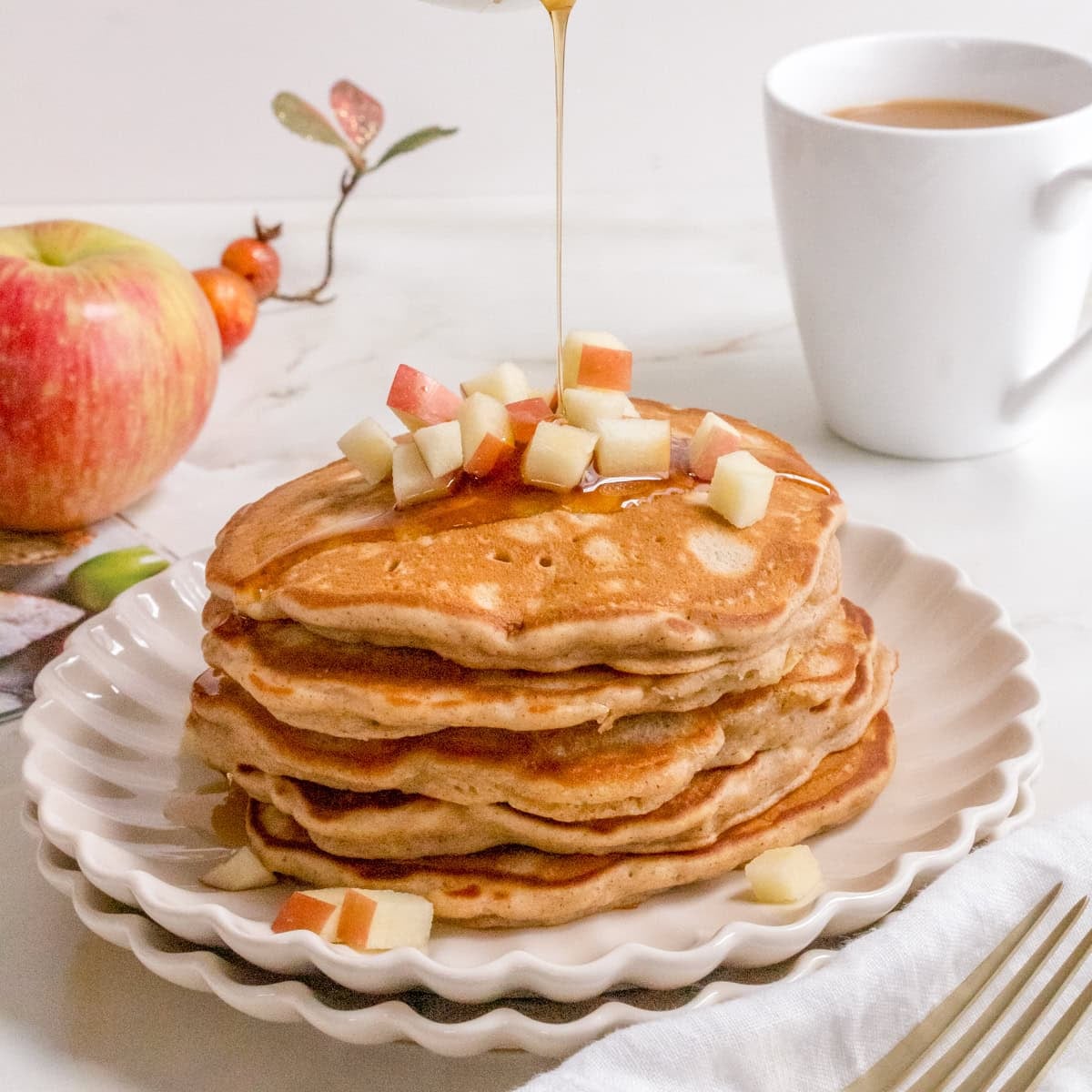 A stack of pancakes topped with diced apples is being drizzled with syrup. A cup of coffee, a fork, and an apple are in the background on a white table.