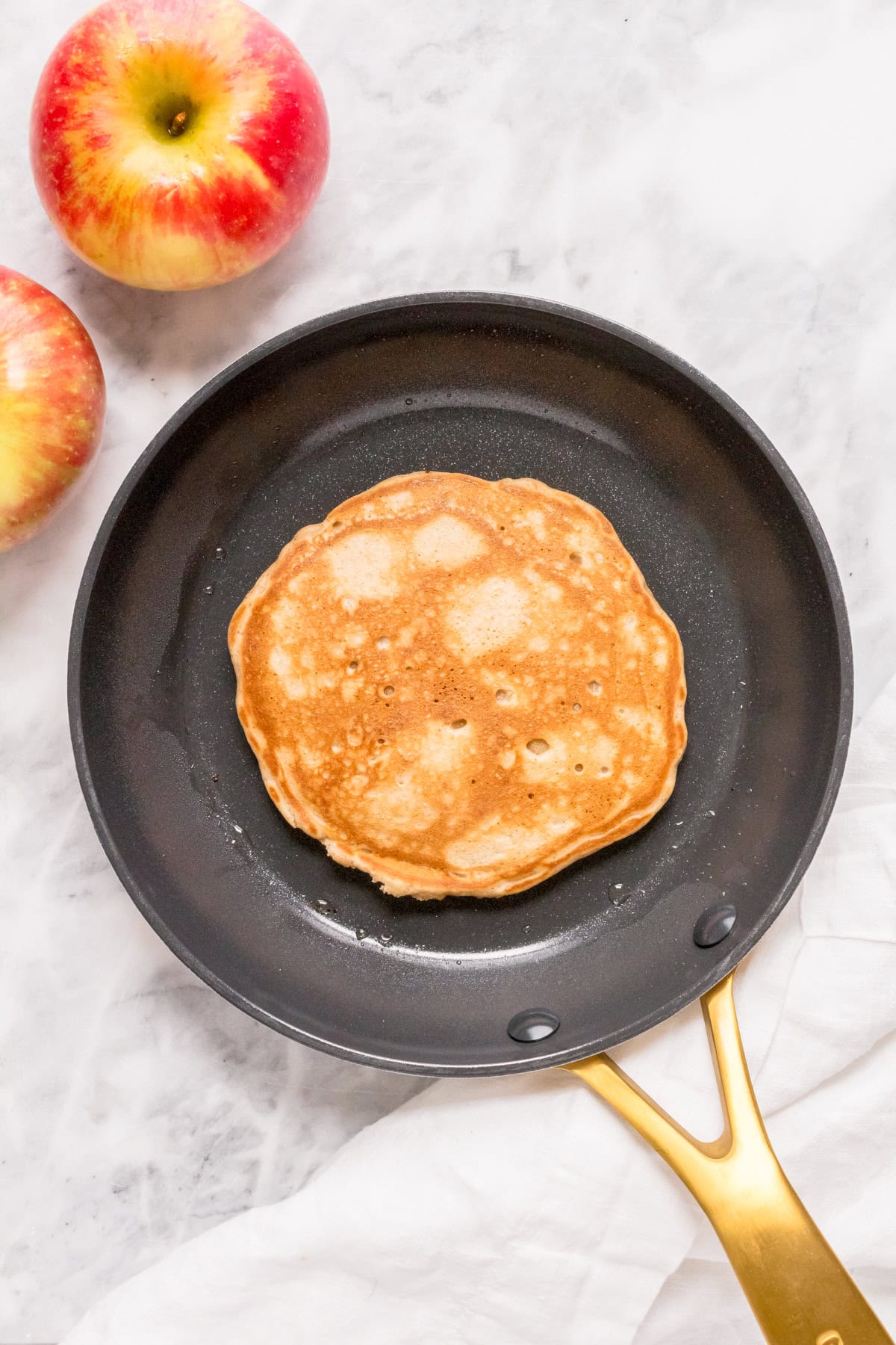 A golden-brown pancake cooking in a black skillet with a gold handle, placed on a marble surface next to two red apples and a white cloth.