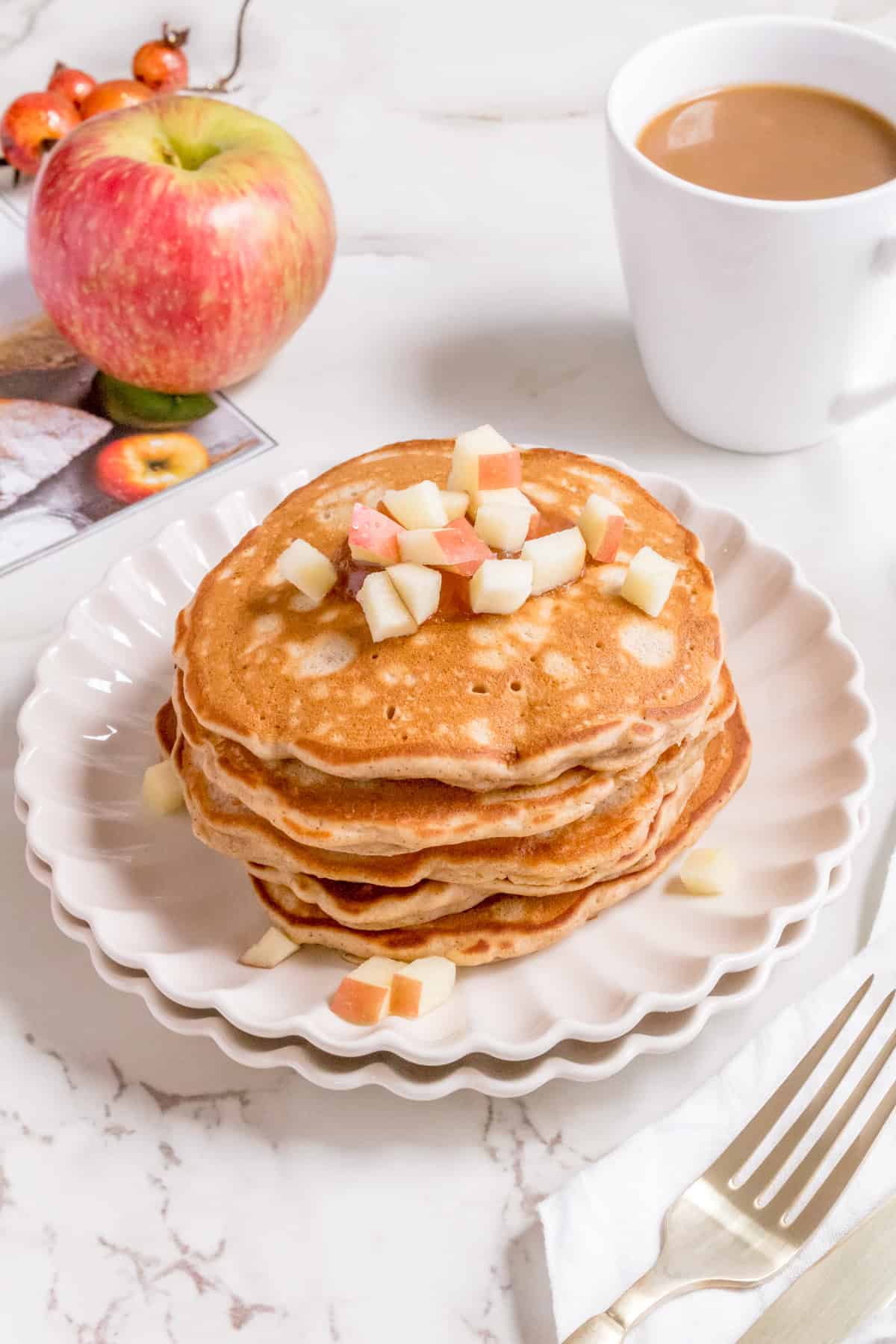 A stack of pancakes topped with diced apples on a white scalloped plate, next to a red apple, a cup of coffee, a fork and knife on a napkin, and a fruit-themed magazine on a marble surface.