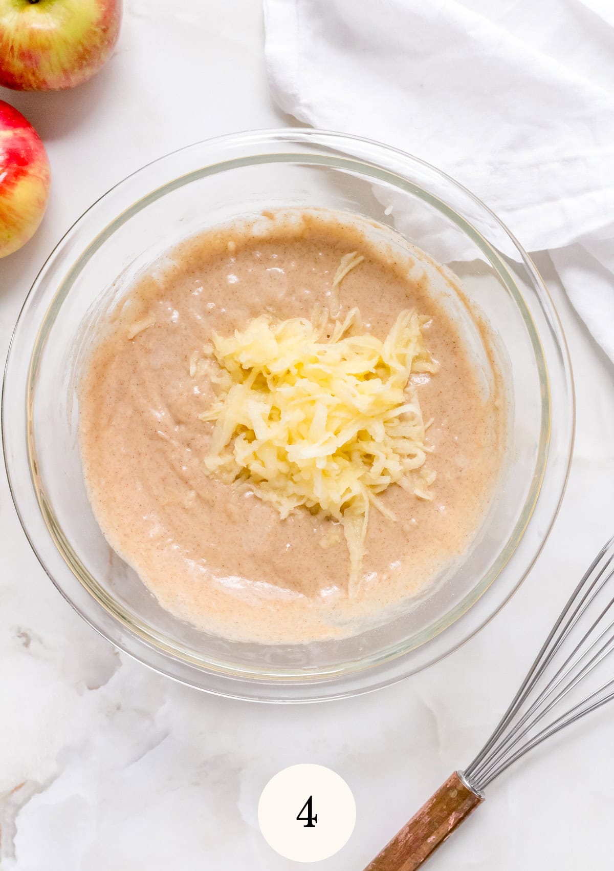 A glass bowl containing batter with grated apple on top sits on a white surface, next to a whisk, apples, and a white cloth. The number 4 is displayed at the bottom.