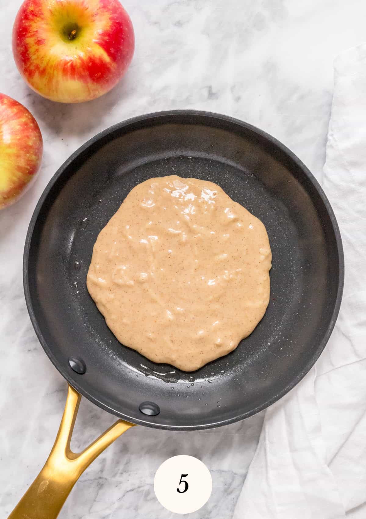 Batter spread in a nonstick skillet with a gold handle, ready to cook; two apples and a white towel are nearby on a marble countertop. A circle with the number 5 is placed at the bottom of the image.