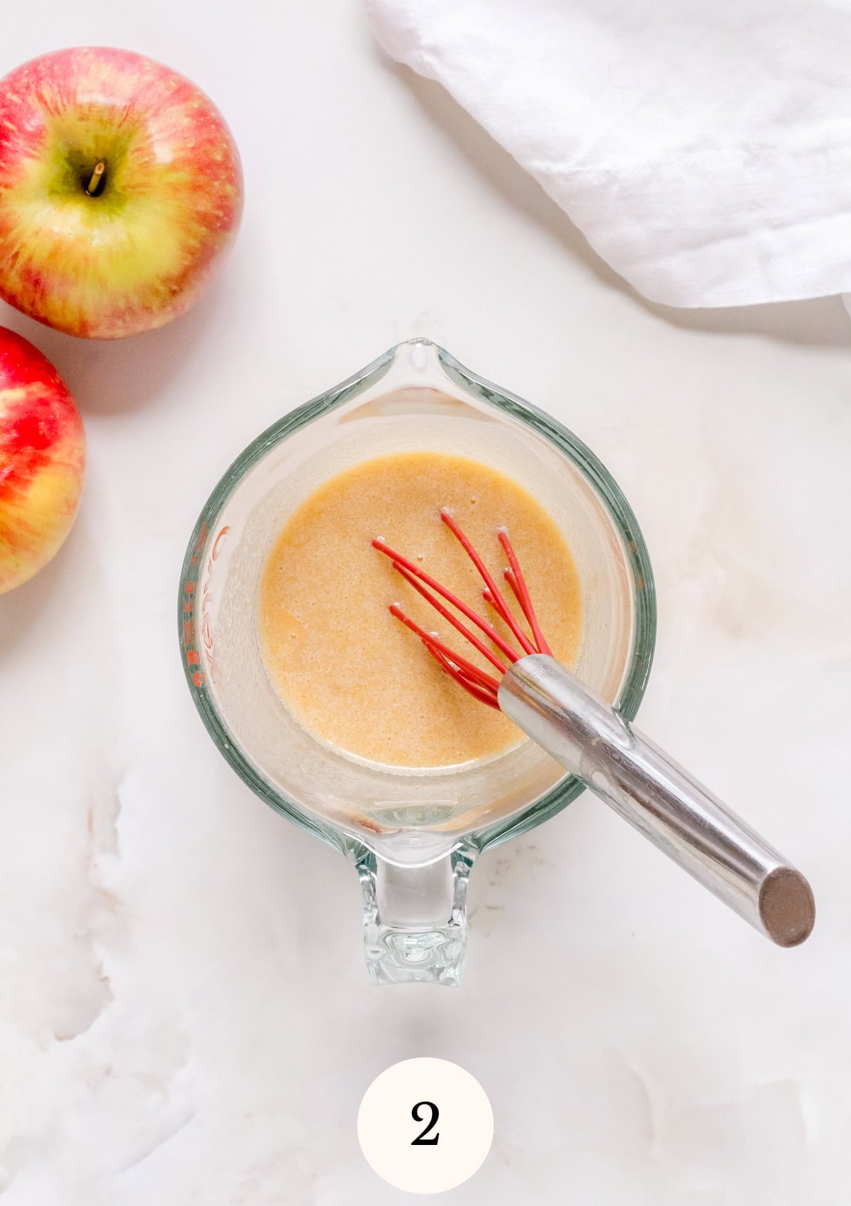 A glass measuring cup with a light brown mixture and a red whisk inside is on a white surface, next to two apples. A white cloth is in the top right corner. The number 2 is at the bottom center.