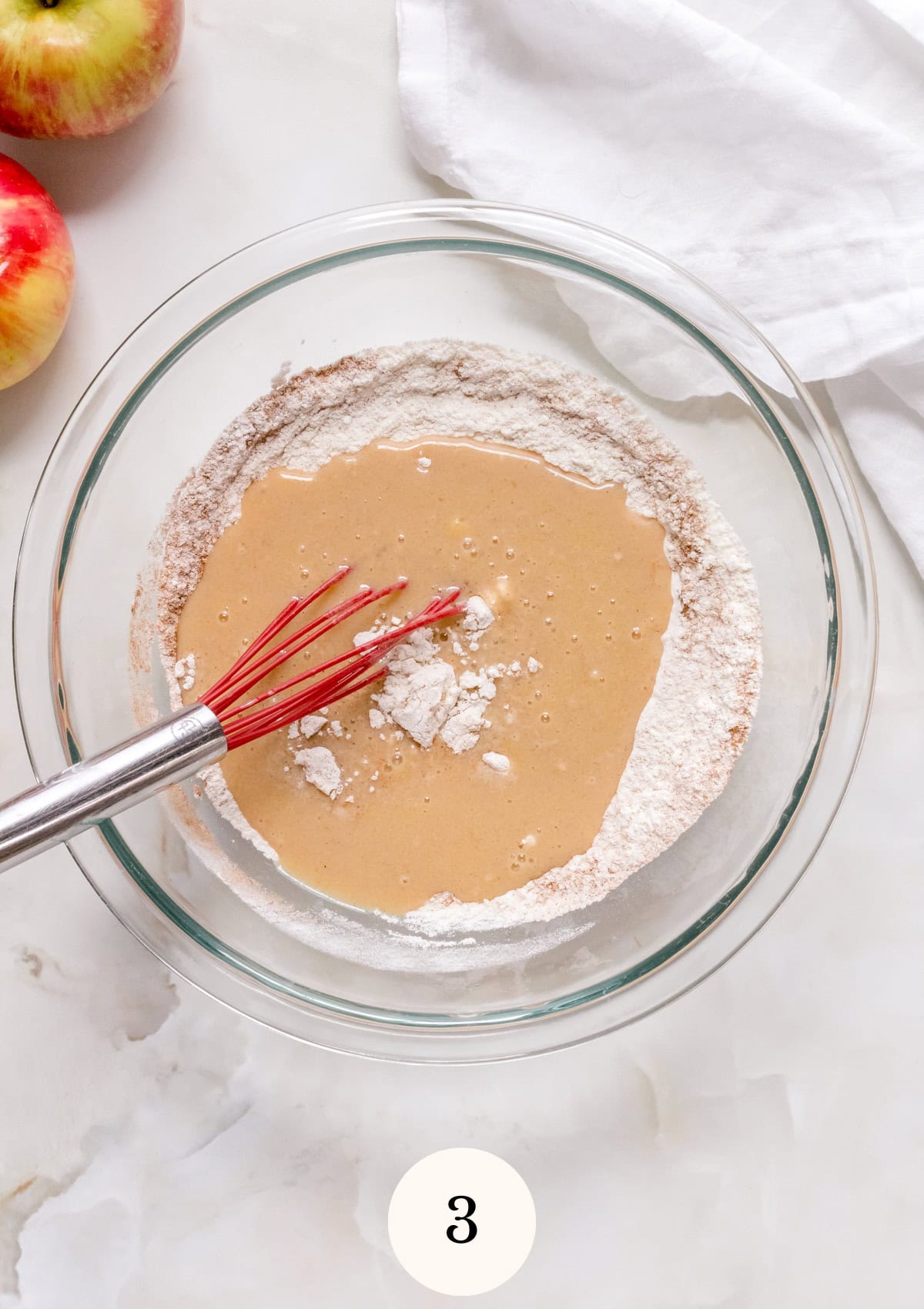 A glass mixing bowl with flour and wet ingredients partially mixed, a red whisk inside. Two apples and a white cloth are nearby on a white surface. The number 3 is shown at the bottom of the image.