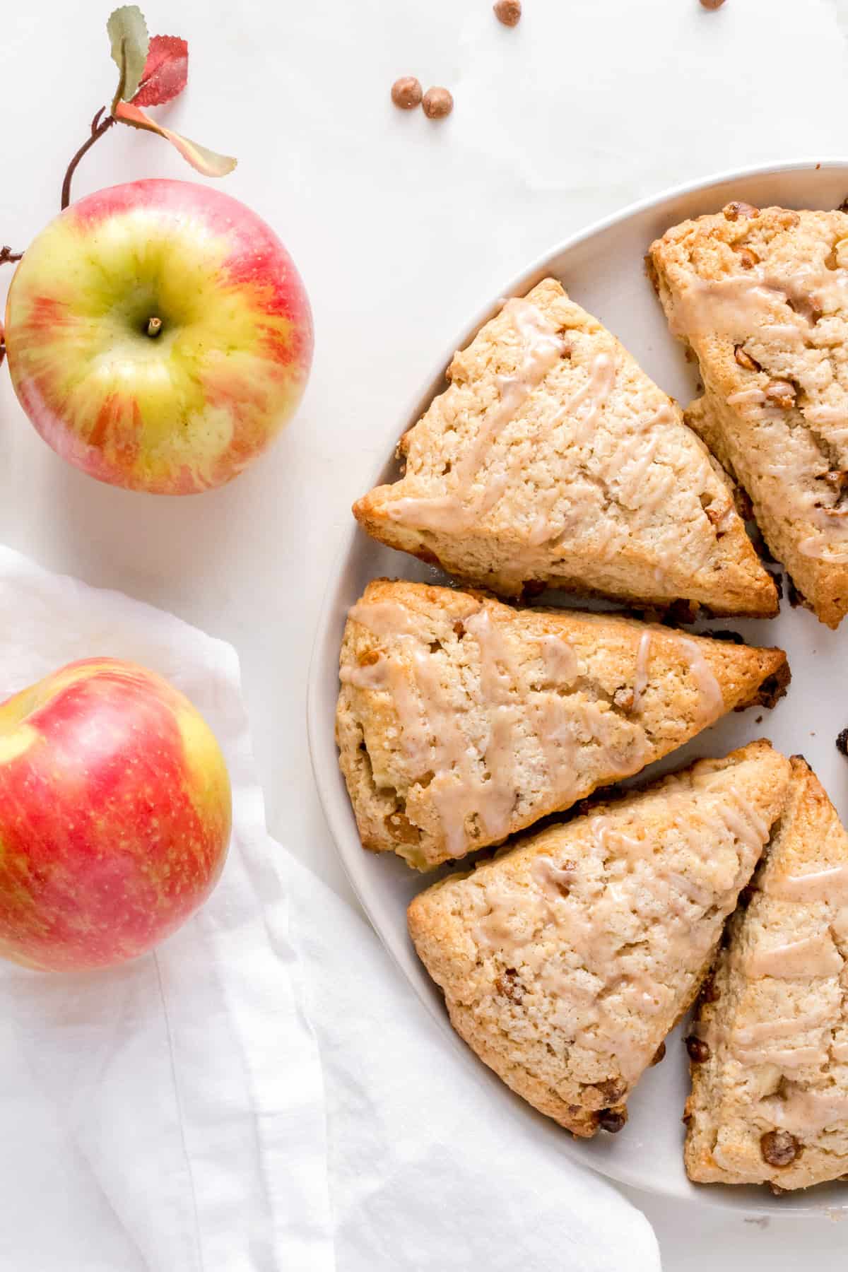 A plate of glazed scones arranged in a circle, with two red and yellow apples and a white cloth nearby on a light surface.