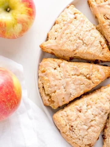 Two red and yellow apples next to a white plate with three glazed scones on a white surface, with part of a white cloth napkin visible.