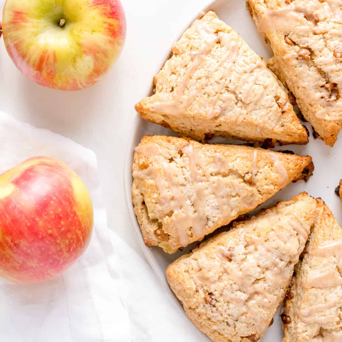 Two red and yellow apples next to a white plate with three glazed scones on a white surface, with part of a white cloth napkin visible.