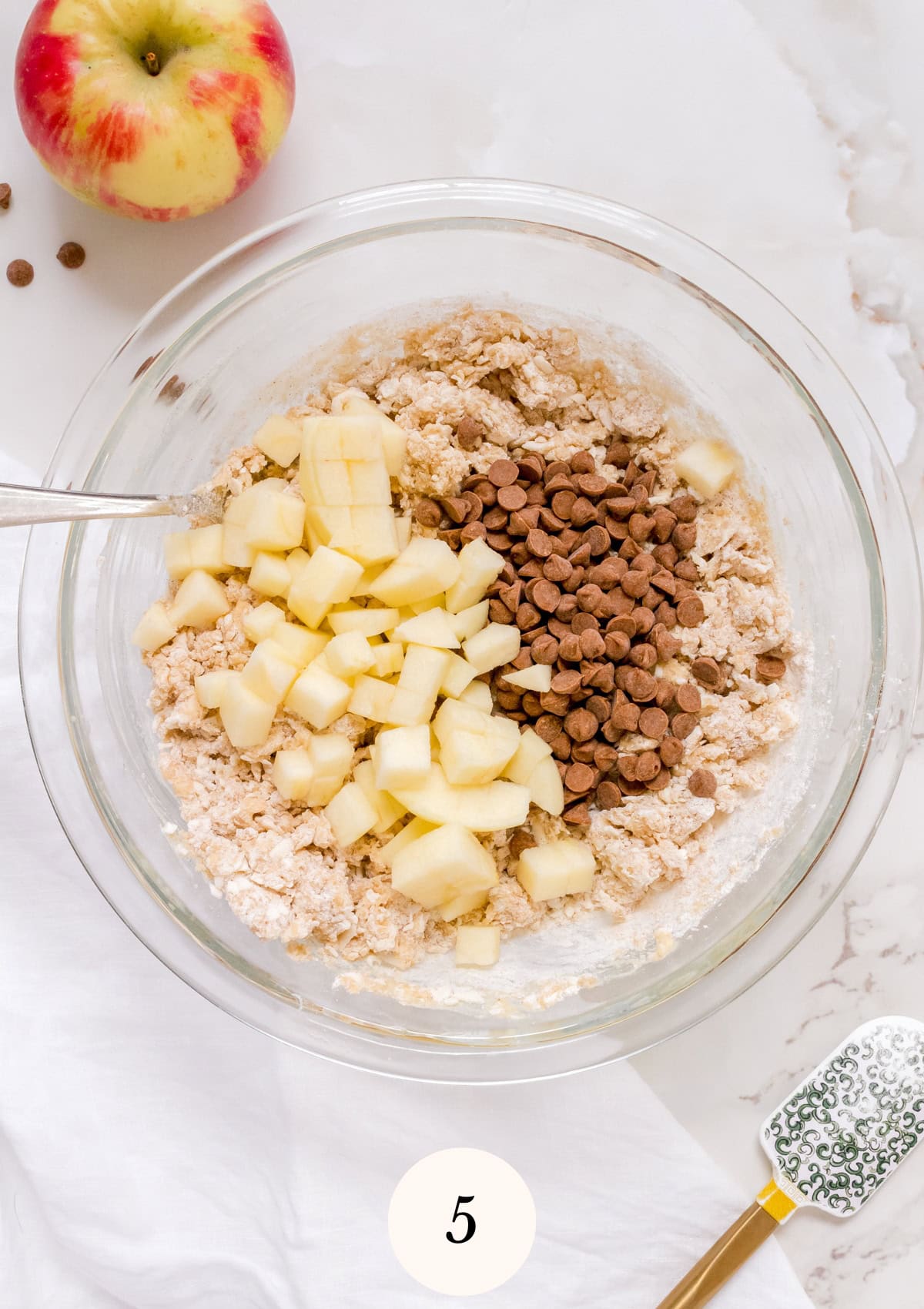 A mixing bowl containing flour mixture, chopped apples, and chocolate chips sits on a white surface. An apple, spatula, and measuring spoon are nearby. The number 5 is shown at the bottom of the image.