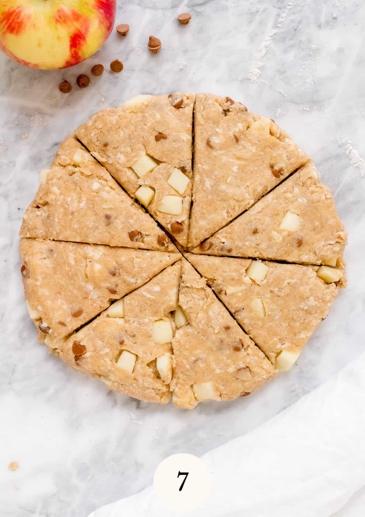 Round, unbaked scone dough with apple chunks and chips, cut into eight wedges on a marble surface. An apple and a few scattered chips are in the top left corner. A white cloth is partially visible at the bottom.