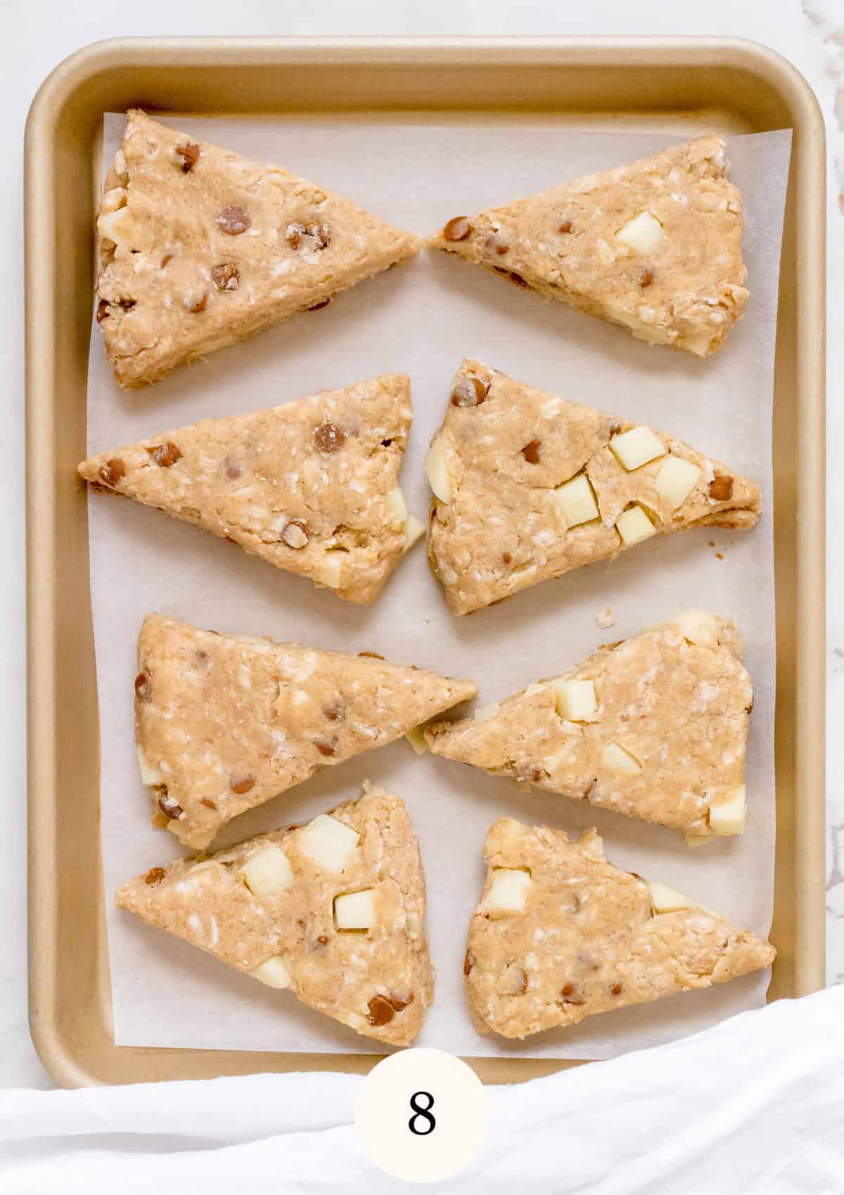 A baking sheet lined with parchment paper holds eight unbaked, triangular scones with visible chunks of nuts and white chocolate. The number 8 is displayed at the bottom of the image.