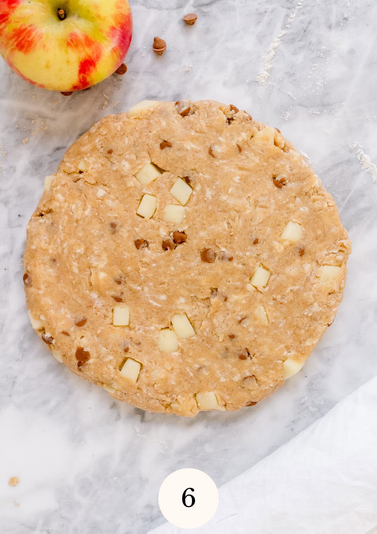 A round disk of unbaked dough with visible apple chunks and nuts sits on a marble surface. An apple is in the upper left corner, and the number 6 is at the bottom center.
