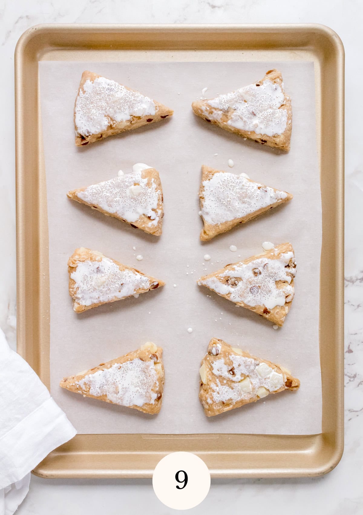 A gold baking tray lined with parchment paper holds nine triangular scones topped with white icing and powdered sugar. The tray rests on a white marble surface, with a cloth in the corner and a card showing the number 9.