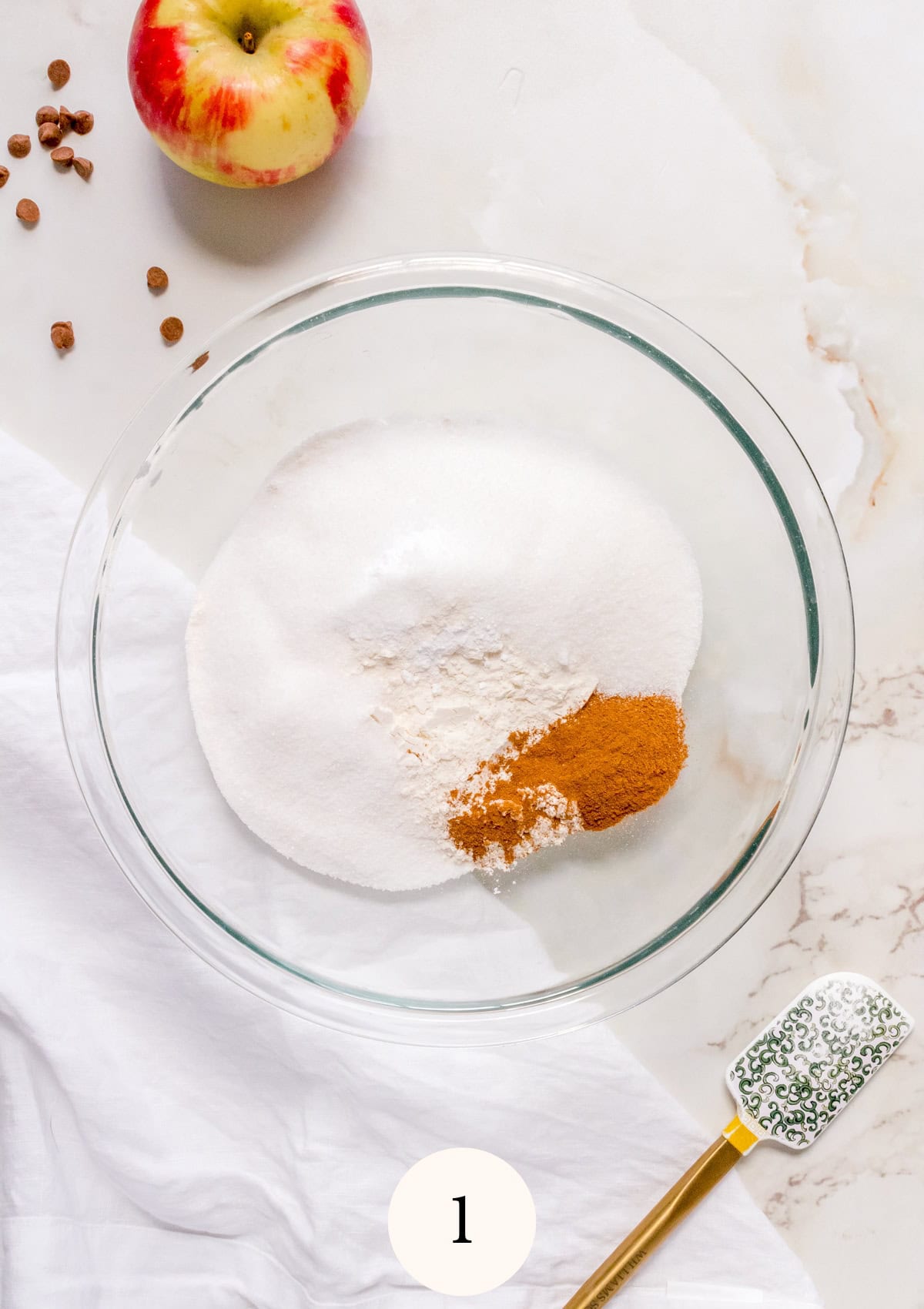 A glass bowl filled with sugar, flour, and cinnamon sits on a marble surface next to a whole apple, scattered cloves, a white cloth, and a spatula with a green patterned handle.