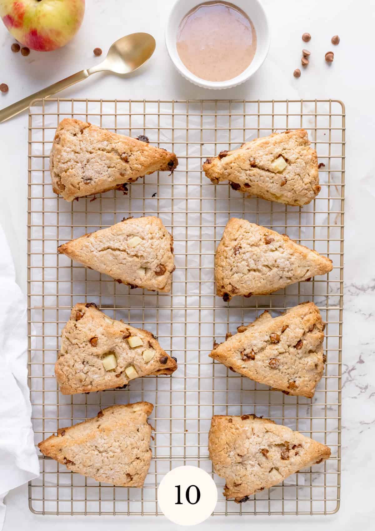 Eight triangle-shaped scones with visible apple pieces and cinnamon chips rest on a wire cooling rack. A bowl of glaze, a spoon, apple, and scattered chips are nearby on a white surface.