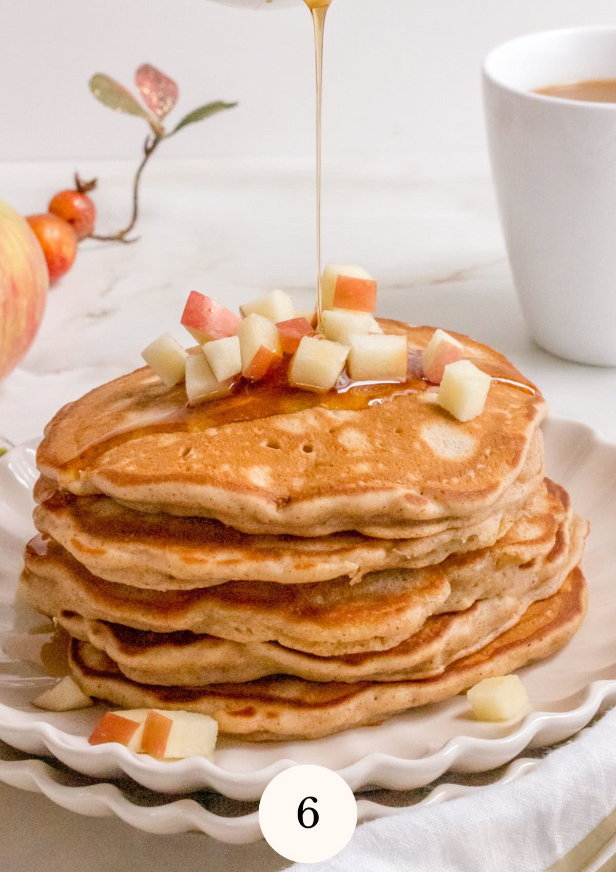 A stack of pancakes topped with diced apples sits on a white plate. Syrup is being poured over the pancakes. In the background, there is a cup of coffee and decorative autumn elements.