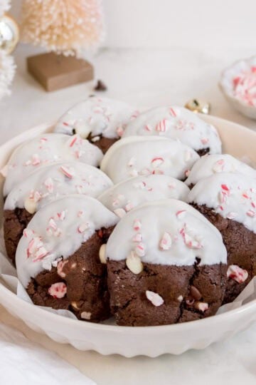 A plate of chocolate cookies partially dipped in white icing and sprinkled with crushed peppermint candy, displayed on a white surface with festive holiday decorations in the background.