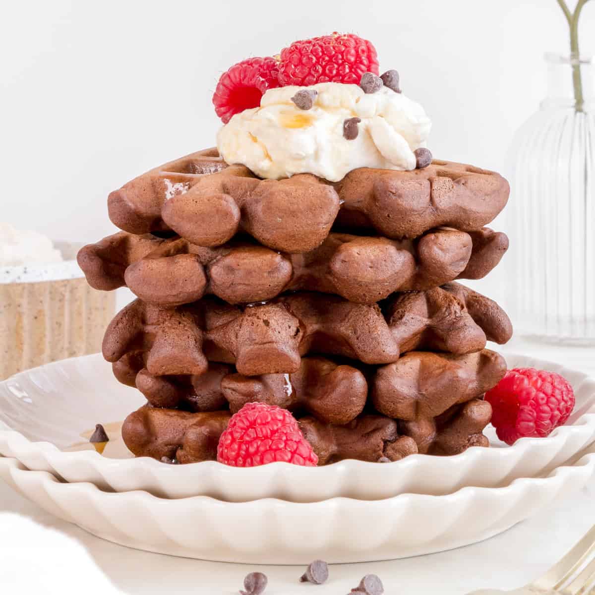 a stack of chocolate waffles with whipped cream and raspberries on a white plate.