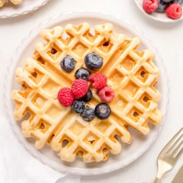 A stack of golden buttermilk waffles on a white plate topped with berries and maple syrup.
