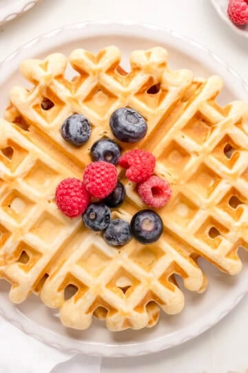 A stack of golden buttermilk waffles on a white plate topped with berries and maple syrup.