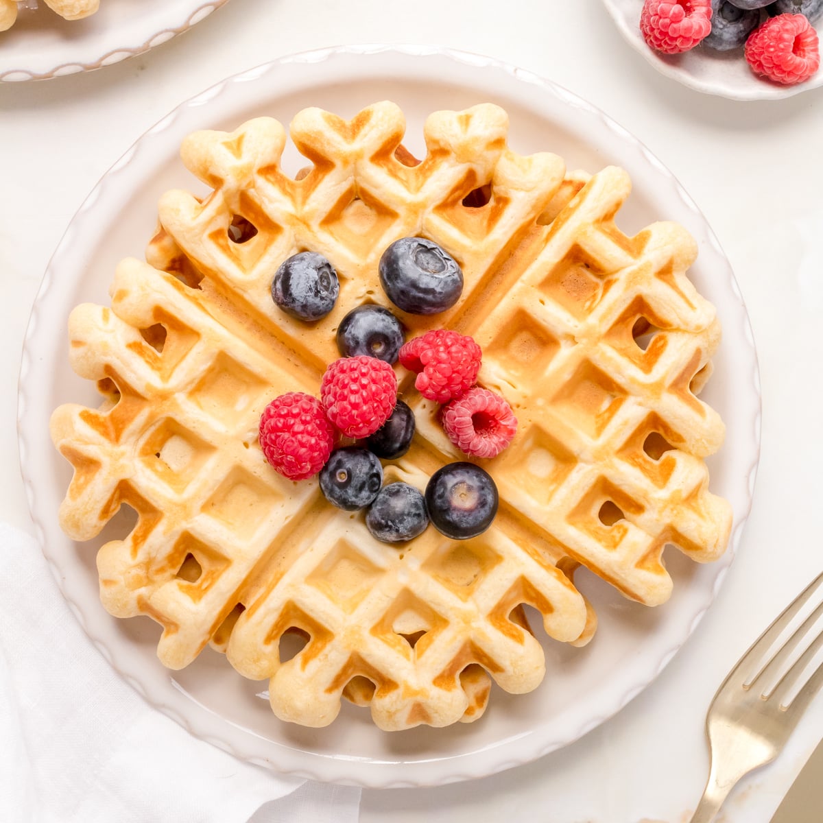 A stack of golden buttermilk waffles on a white plate topped with berries and maple syrup.