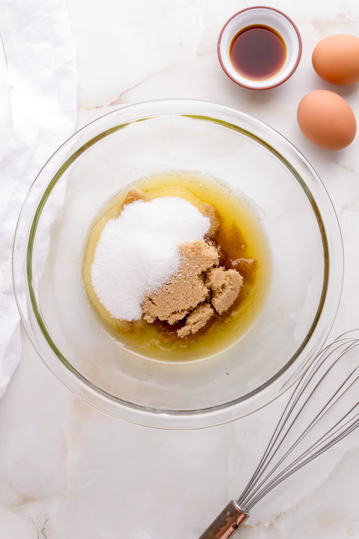 white and brown sugar added to brown butter on white counter with eggs and whisk.