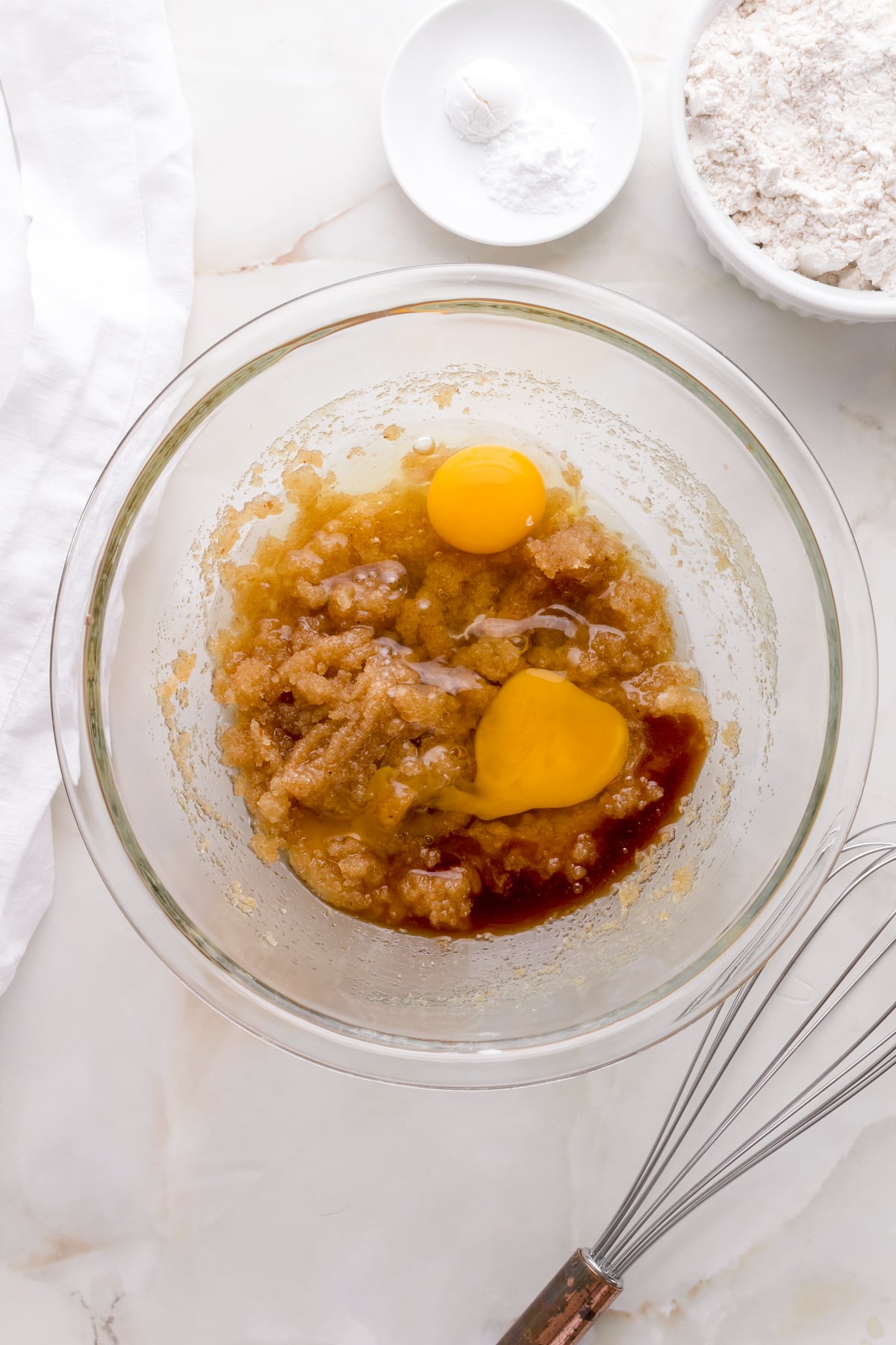 adding eggs to gluten free chocolate chip cookie dough on white counter with copper whisk.
