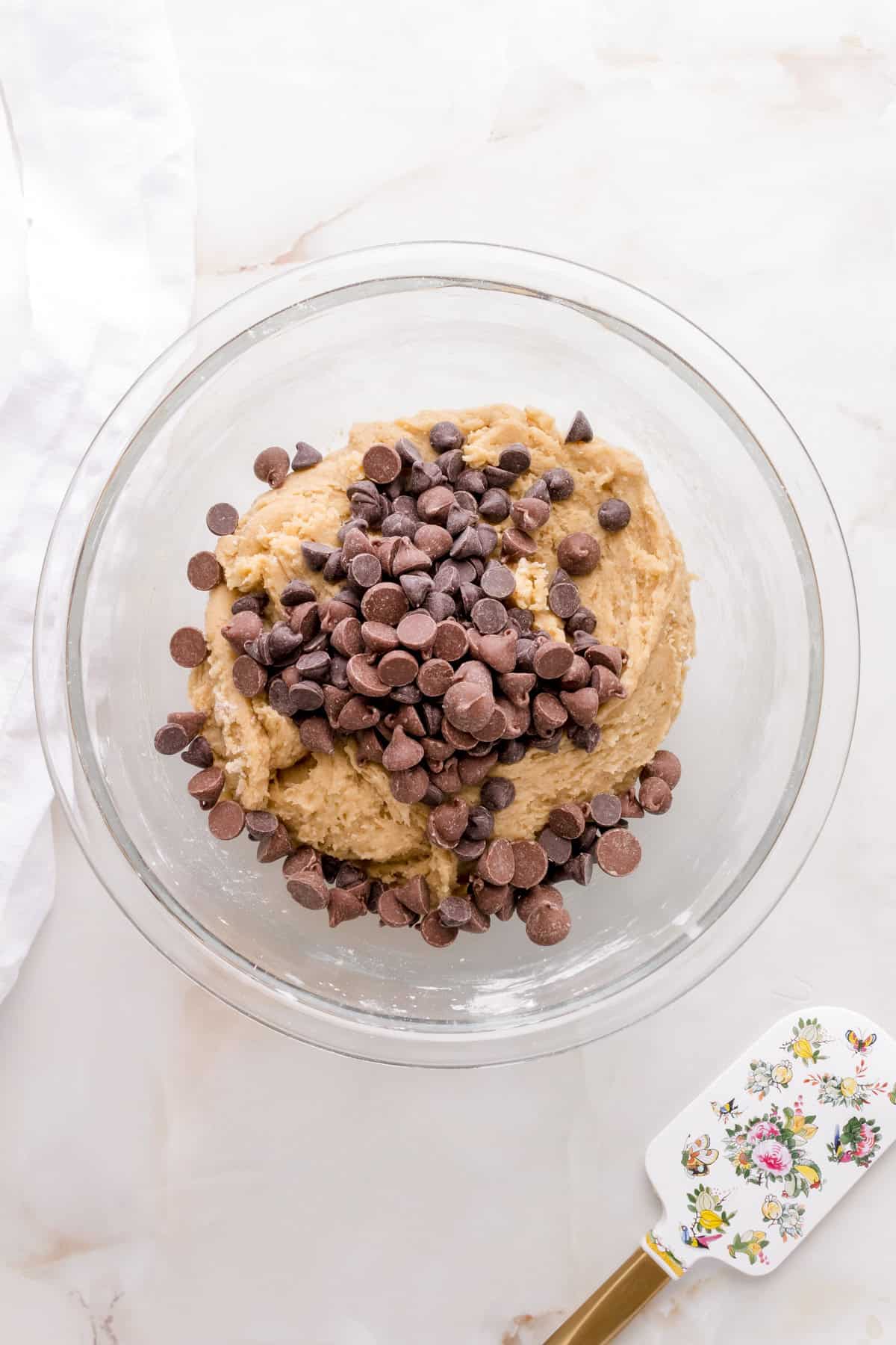 adding chocolate chip to gluten free cookie dough on white counter with spatula.