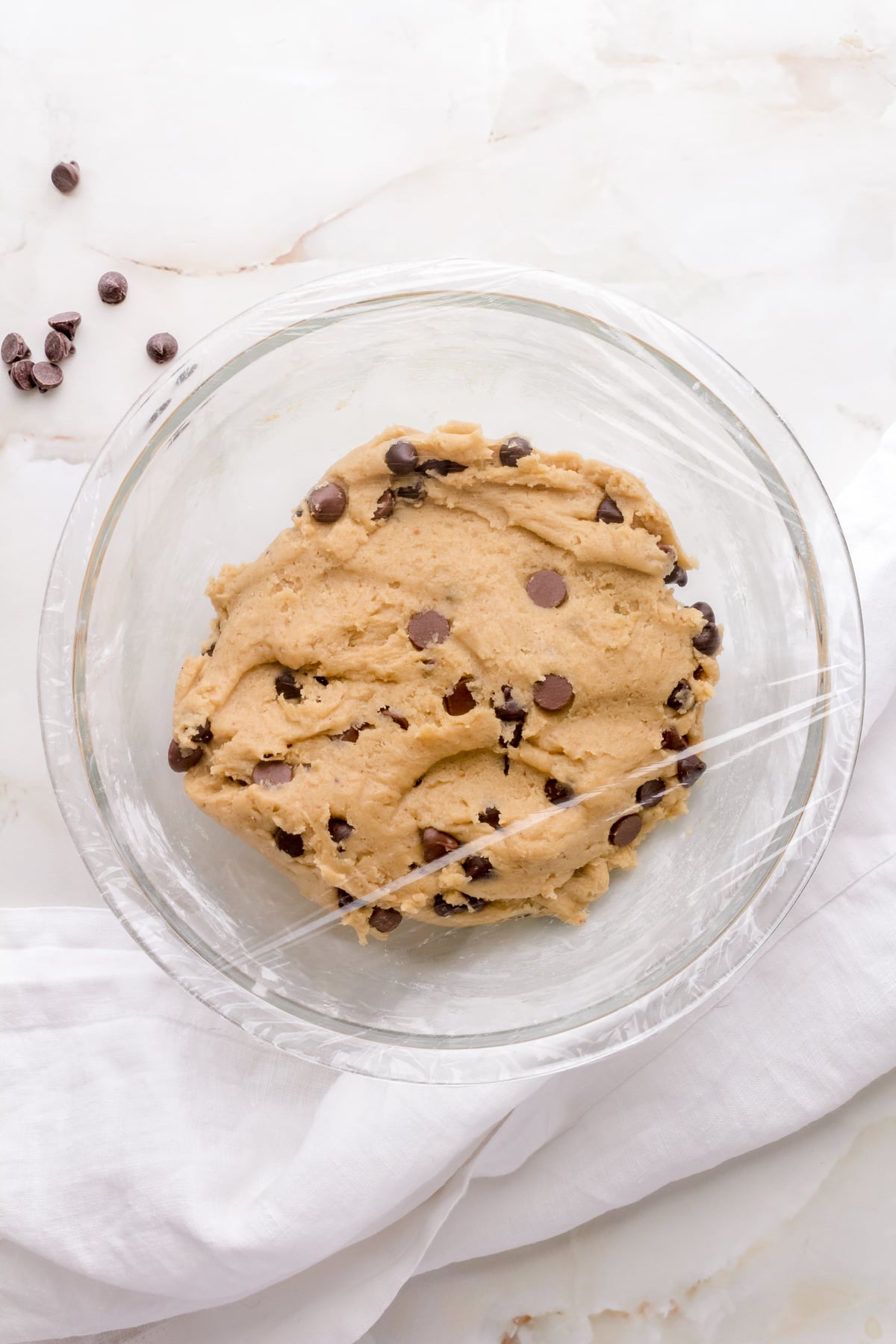 covered bowl of gluten free chocolate chip cookie dough on white counter with white napkin.