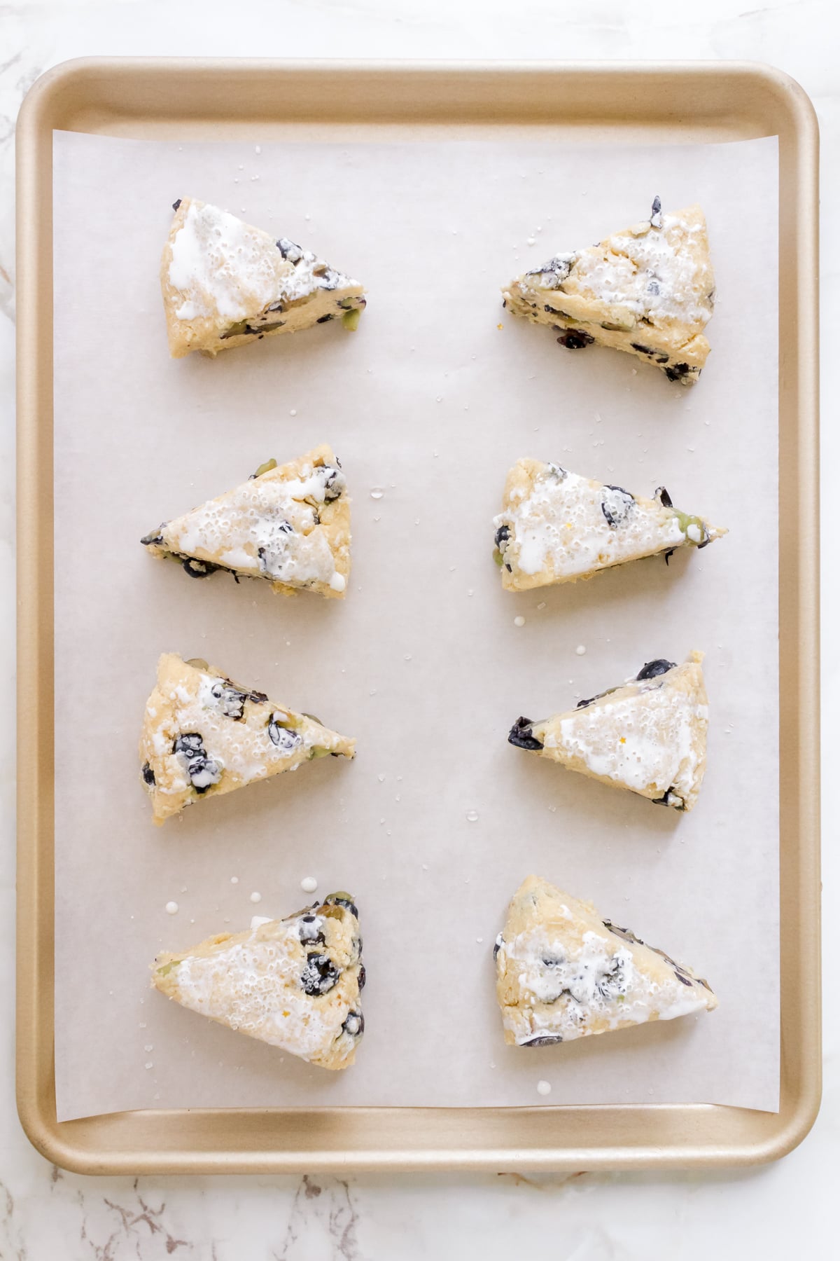 blueberry scones brushed with heavy cream on parchment lined gold sheet pan.
